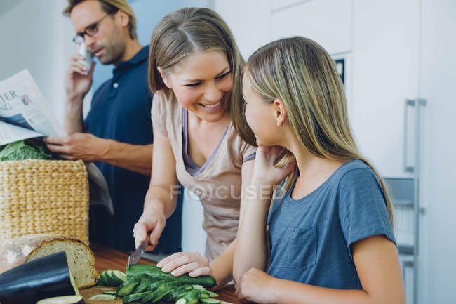 Mother and daughter in kitchen slicing cucumber with father on phone in background — Stock Photo