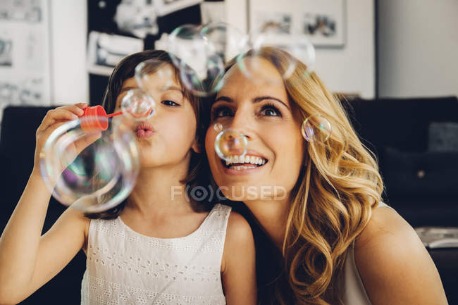 Mère heureuse avec sa fille à la maison soufflant des bulles de savon — Photo de stock