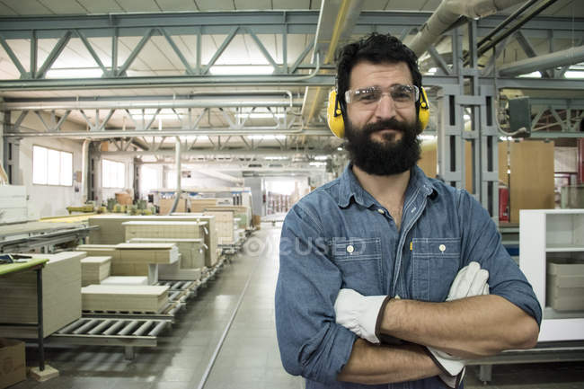 Человек со слуховой защитой, защитными очками и перчатками улыбается, скрестив руки на заводе. — стоковое фото