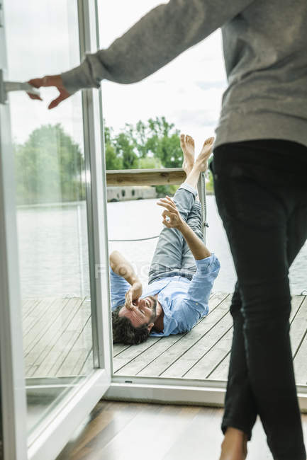 Uomo sdraiato sul ponte di legno di una casa barca con donna che apre la porta del balcone — Foto stock