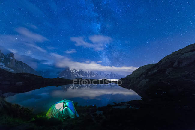 Vista panorámica de la carpa iluminada en la orilla del lago por la noche con la Vía Láctea y el Monte Blanc reflejado en el lago, Lago Cheserys, Mont Blanc, Francia - foto de stock