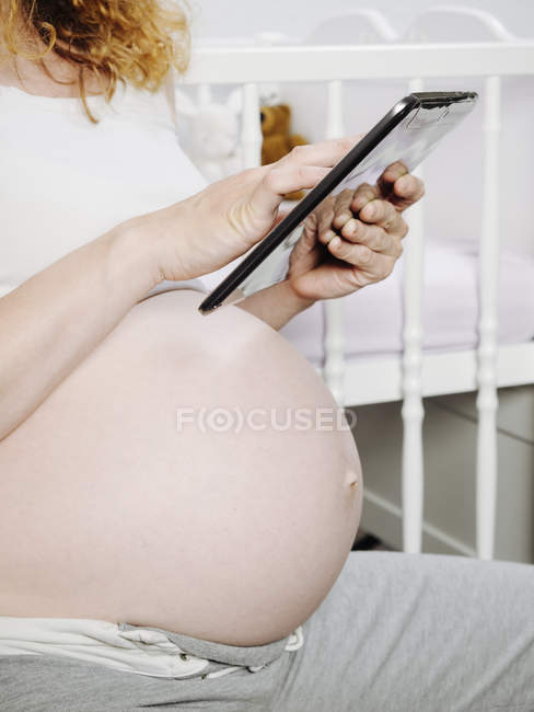 Беременная женщина перед детской кроваткой с помощью цифрового планшета — стоковое фото