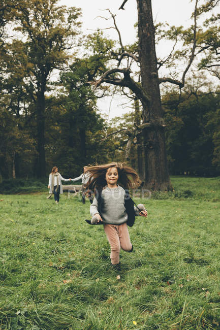 Petite fille courant sur une prairie dans un parc tandis que ses parents se tiennent en arrière-plan — Photo de stock