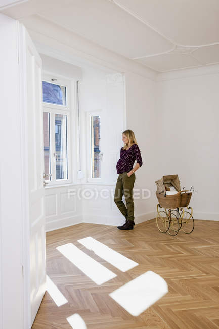 Young pregnant woman with pram in an empty room looking out of window — Stock Photo