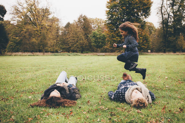 Padres jóvenes tumbados en un prado mientras una niña salta sobre ellos en un parque - foto de stock
