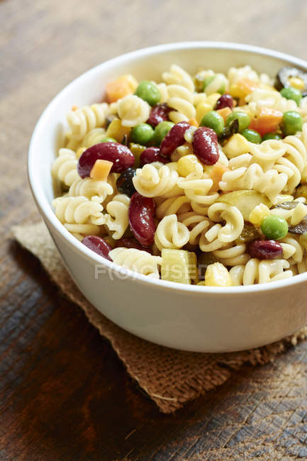 Pasta salad with fusilli pasta, kidney beans, corn, carrots, peas, pickles and black olives served in white bowl — Stock Photo