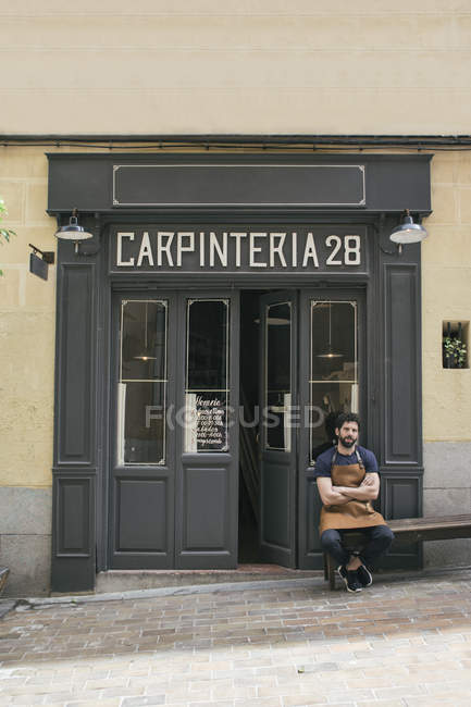 Handwerker mit Lederschürze und verschränkten Armen sitzt am Eingang seiner Werkstatt — Stockfoto