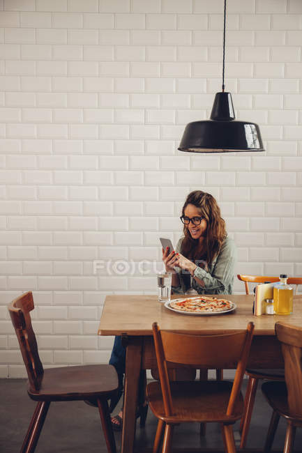 Mujer joven comiendo pizza en el restaurante, usando el teléfono móvil - foto de stock