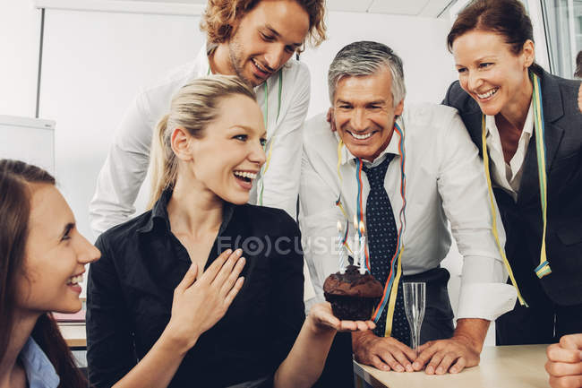 Empresaria emocionada sosteniendo panecillo pequeño con velas encendidas celebrando cumpleaños en la oficina con colegas - foto de stock