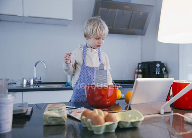 Niño pequeño de pie en la cocina hornear con la ayuda de la tableta digital - foto de stock