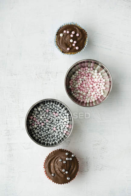 Two cupcakes with chocolate cream and sugar pearls and two bowls of sugar pearls — Stock Photo