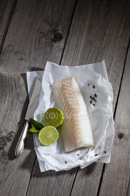 Filete de bacalao, lima y cuchillo, y granos de pimienta sobre papel a prueba de grasa sobre madera - foto de stock