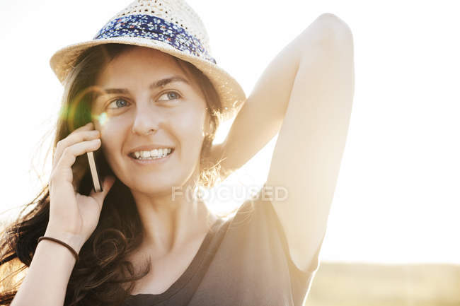 Mujer sonriente que usa sombrero de verano telefoneando con teléfono inteligente - foto de stock