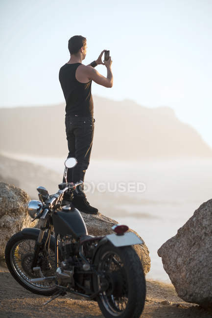 Ciudad del Cabo, Sudáfrica, motociclista de pie en la roca en la costa tomando fotos - foto de stock