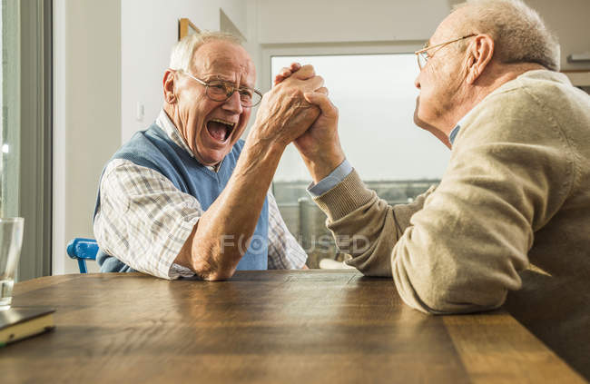 Two Senior Friends Arm Wrestling At Home Side View