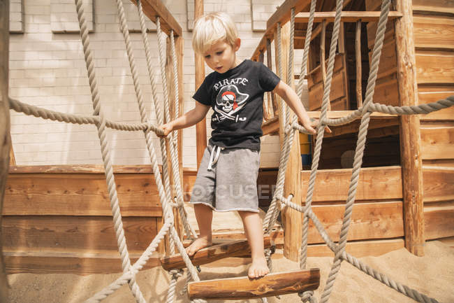 Petit garçon marchant le long du pont suspendu du bateau en bois sur l'aire de jeux — Photo de stock