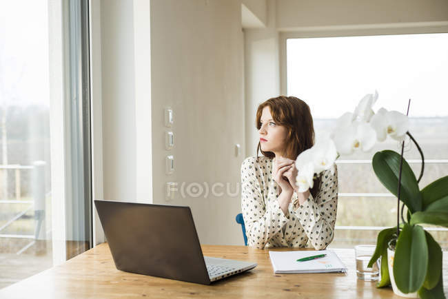 Giovane donna con computer portatile al tavolo di legno guardando fuori dalla finestra — Foto stock