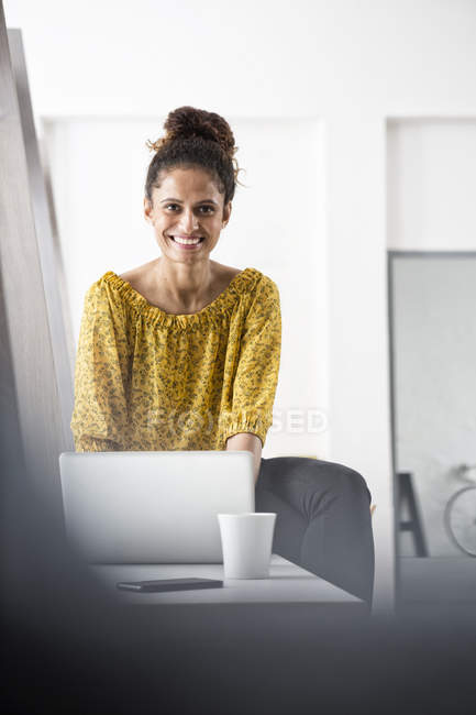 Mujer sonriente sentada en el escritorio de la oficina usando computadora portátil - foto de stock
