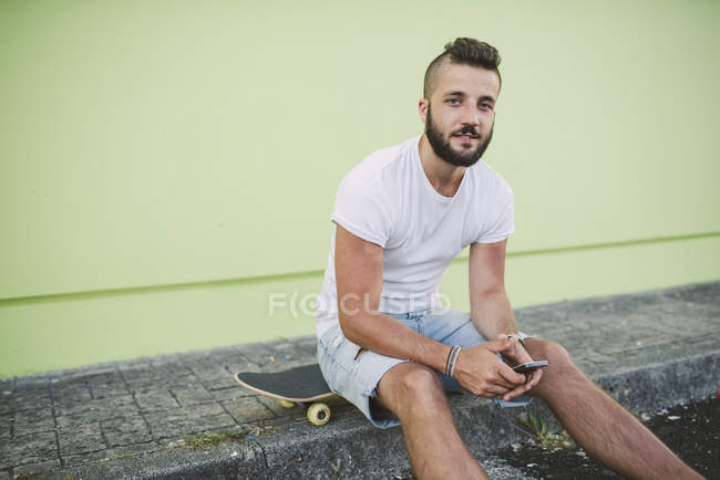 Portrait d'homme avec smartphone assis sur planche à roulettes sur trottoir — Photo de stock