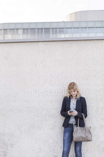 Mulher de negócios loira em pé na frente da parede olhando para o smartphone — Fotografia de Stock
