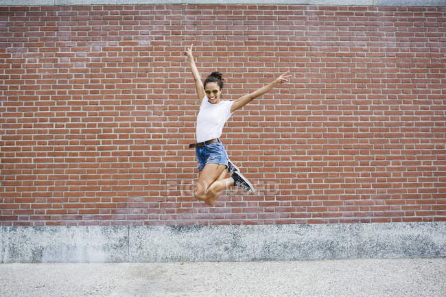 Giovane donna che salta su con le mani in alto, muro di mattoni su sfondo — Foto stock