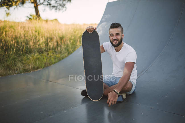 Smiling skateboarder sentado en la rampa en skatepark - foto de stock