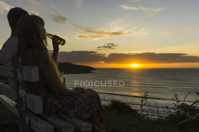 Couple assis sur le banc au coucher du soleil et boire des boissons dans des bouteilles en verre — Photo de stock