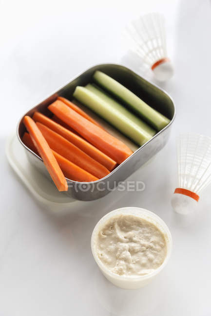 Box of cucumber and carrot sticks and a bowl of dip on white ground — Stock Photo