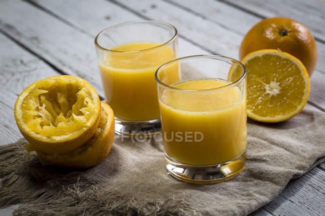 Dos vasos de zumo de naranja recién exprimido y naranjas sobre tela sobre madera - foto de stock