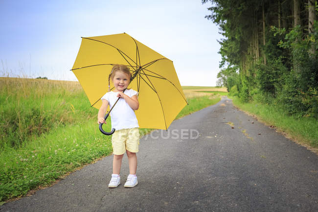 Усміхаючись дівчинка з жовтий парасолька стоїть на дорозі — стокове фото