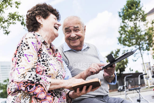 Rindo casal de idosos sentados juntos em um banco com livro e tablet — Fotografia de Stock
