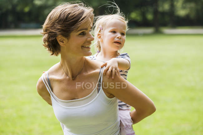 Mother carrying her little daughter piggy back in a park — Stock Photo