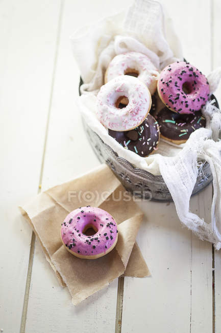 Pink, chocolate and white donuts with sprinkles in basket on white wooden table — Stock Photo