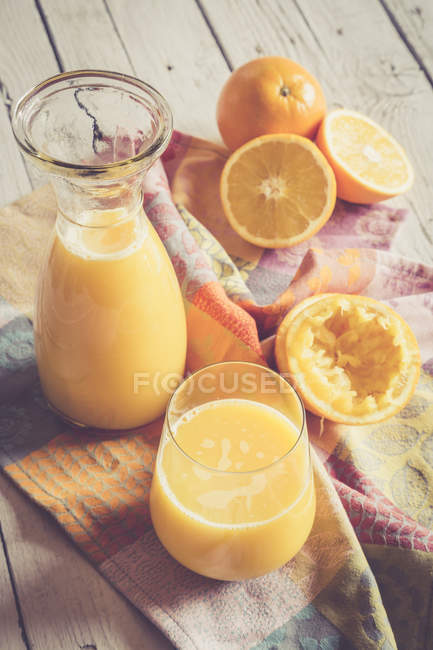 Naranjas frescas con jarra y vaso de jugo de naranja recién exprimido sobre tela sobre madera - foto de stock