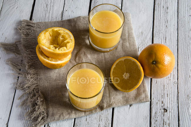 Dos vasos de zumo de naranja recién exprimido y naranjas sobre tela y madera - foto de stock