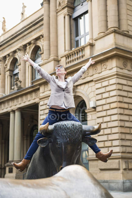 Mujer de negocios con los brazos extendidos sentado en la escultura del toro en frente de la bolsa, Alemania, Hesse, Frankfurt - foto de stock