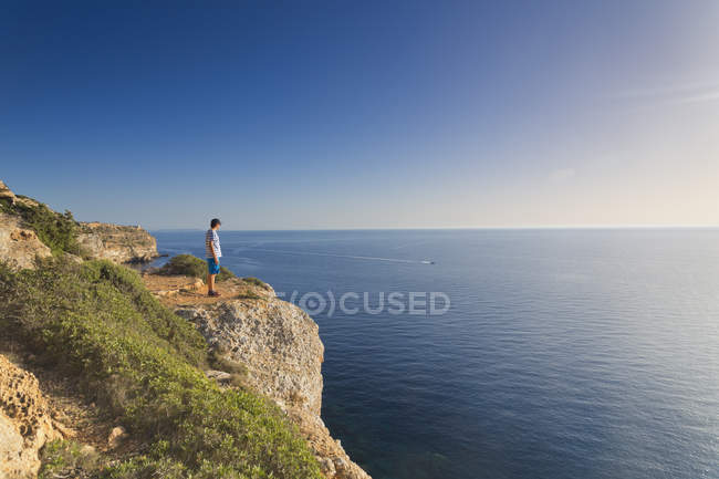 Spain, Balearic Islands, Majorca, one teenage boy standing on a rock at the cliff coast, watching the sundown — Stock Photo