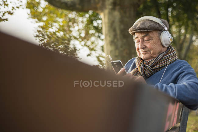Uomo anziano sulla panchina del parco con cellulare e cuffie — Foto stock