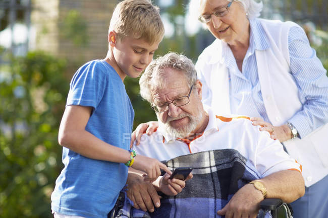 Nonna e nipote spiegano il cellulare al nonno sulla sedia a rotelle — Foto stock