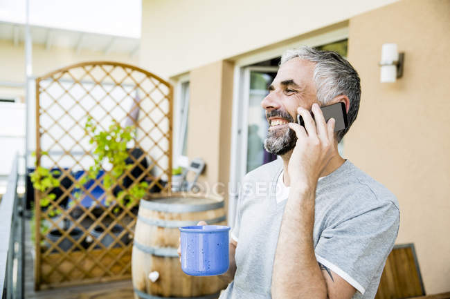 Uomo sul suo balcone telefonare con smartphone in possesso di una tazza di caffè — Foto stock
