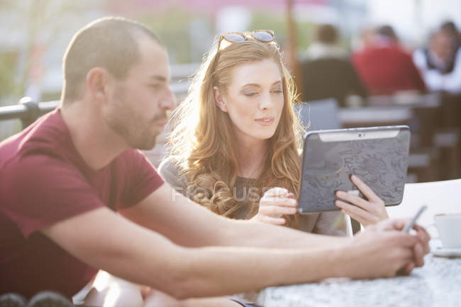 Pareja tomando un descanso de café en la cafetería al aire libre, hombre con teléfono inteligente y mujer sosteniendo tableta digital sentado en la mesa - foto de stock