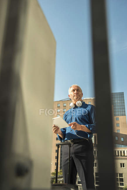 Uomo d'affari con valigia, tablet digitale e cuffie fuori dall'edificio per uffici — Foto stock