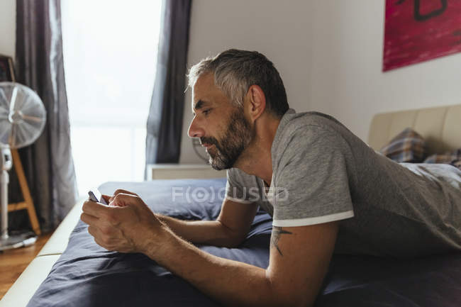 Мужчина лежит на кровати со своим смартфоном — стоковое фото