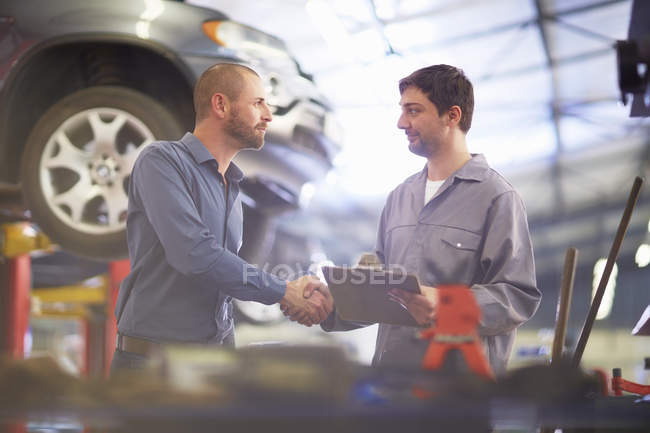 Meccanico auto e cliente stringendo la mano in garage di riparazione, auto sullo sfondo — Foto stock