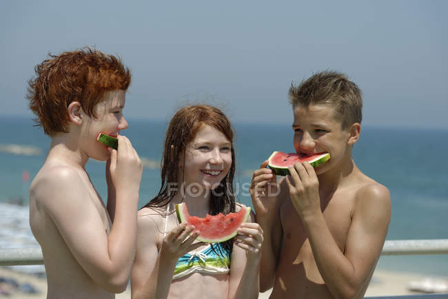 Chica y dos adolescentes comiendo rodajas de sandía en la playa - foto de stock
