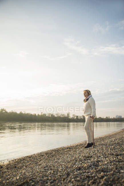 Senior woman with walking stick standing at waterside watching sunset — Stock Photo