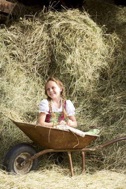 Bonito jovem menina no tradicional dirndl sentado no carrinho de mão no feno — Fotografia de Stock