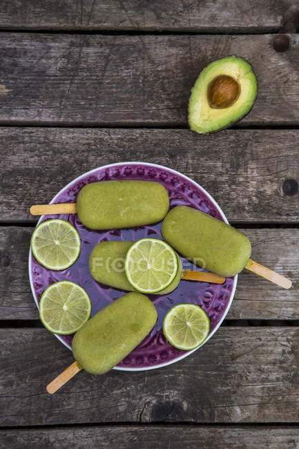 Elevated view of plate with avocado ice lollies and slices of lime on wooden surface — Stock Photo