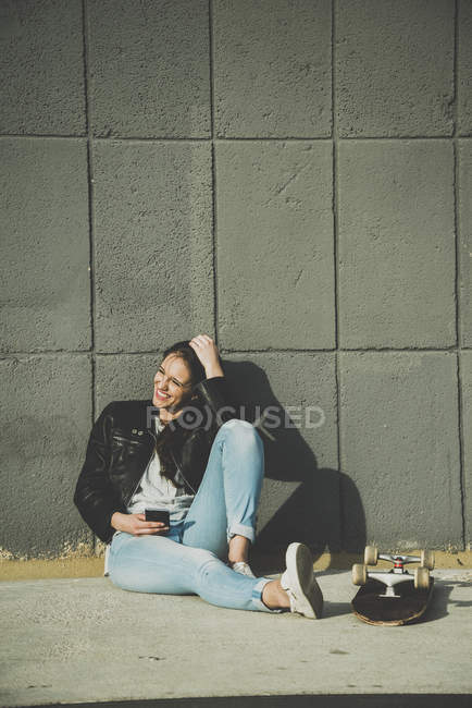 Молодая женщина сидит на улице со скейтбордом и сотовым телефоном — стоковое фото