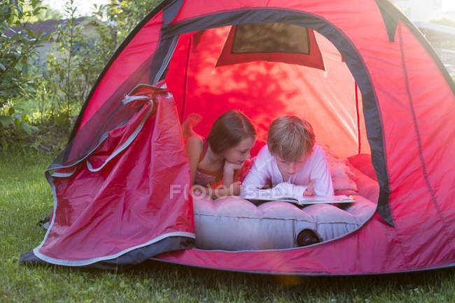 Niño y niña leyendo acostados en una carpa roja con un libro - foto de stock